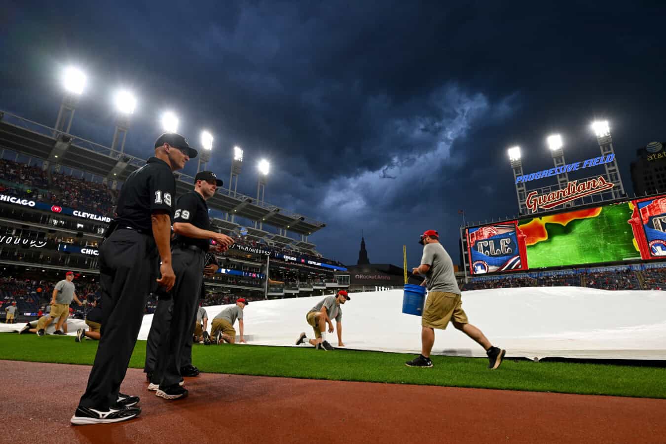 CLEVELAND, OHIO - JULY 03: Umpire Vic Carapazza #19 and umpire Nic Lentz #59 watch as members of the Cleveland Guardians grounds crew secure the tarp on the field during a rain delay during the fifth inning of the game between the Cleveland Guardians and the Chicago White Sox at Progressive Field on July 03, 2026 in Cleveland, Ohio.